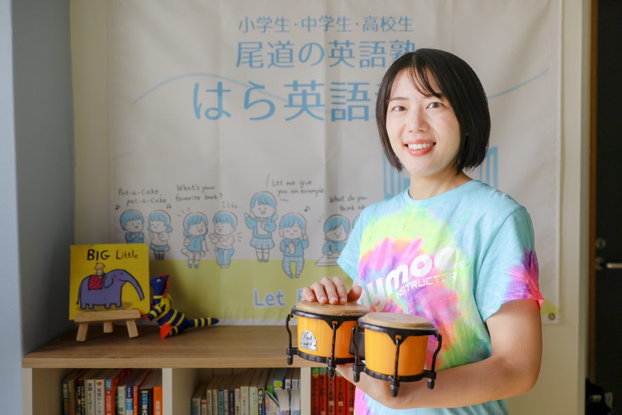 教室の看板の前で、楽器を持ってほほえむ講師 原沙織